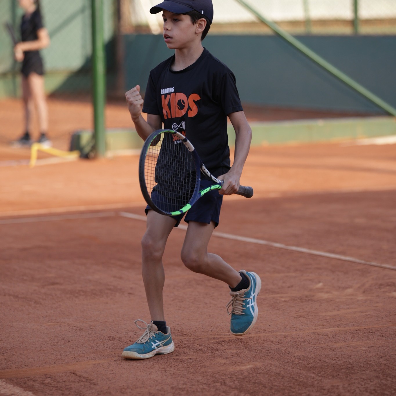 Aluno kids correndo na quadra durante aula de tennis.