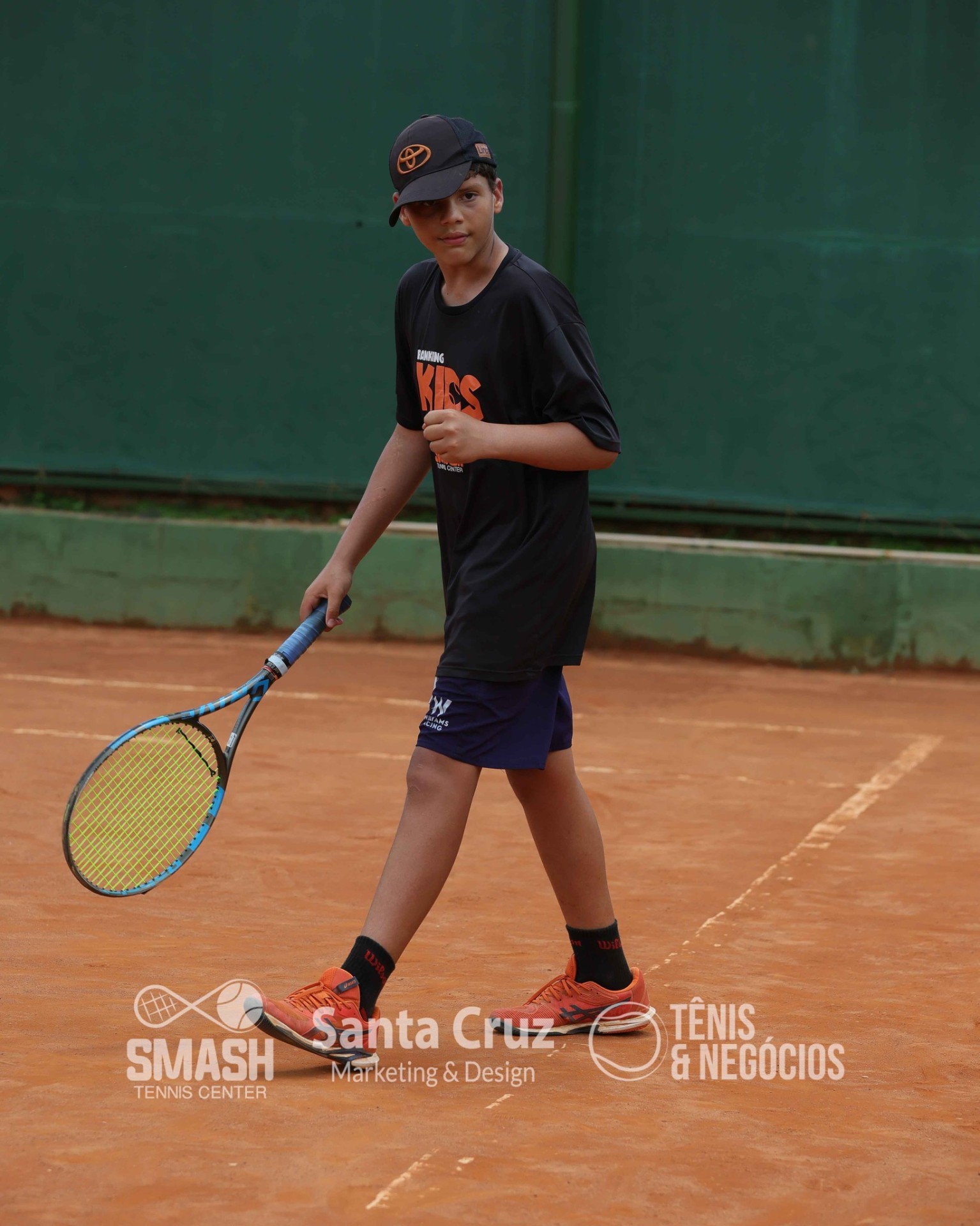 Aluno kids com raquete na mão em quadra de saibro.
